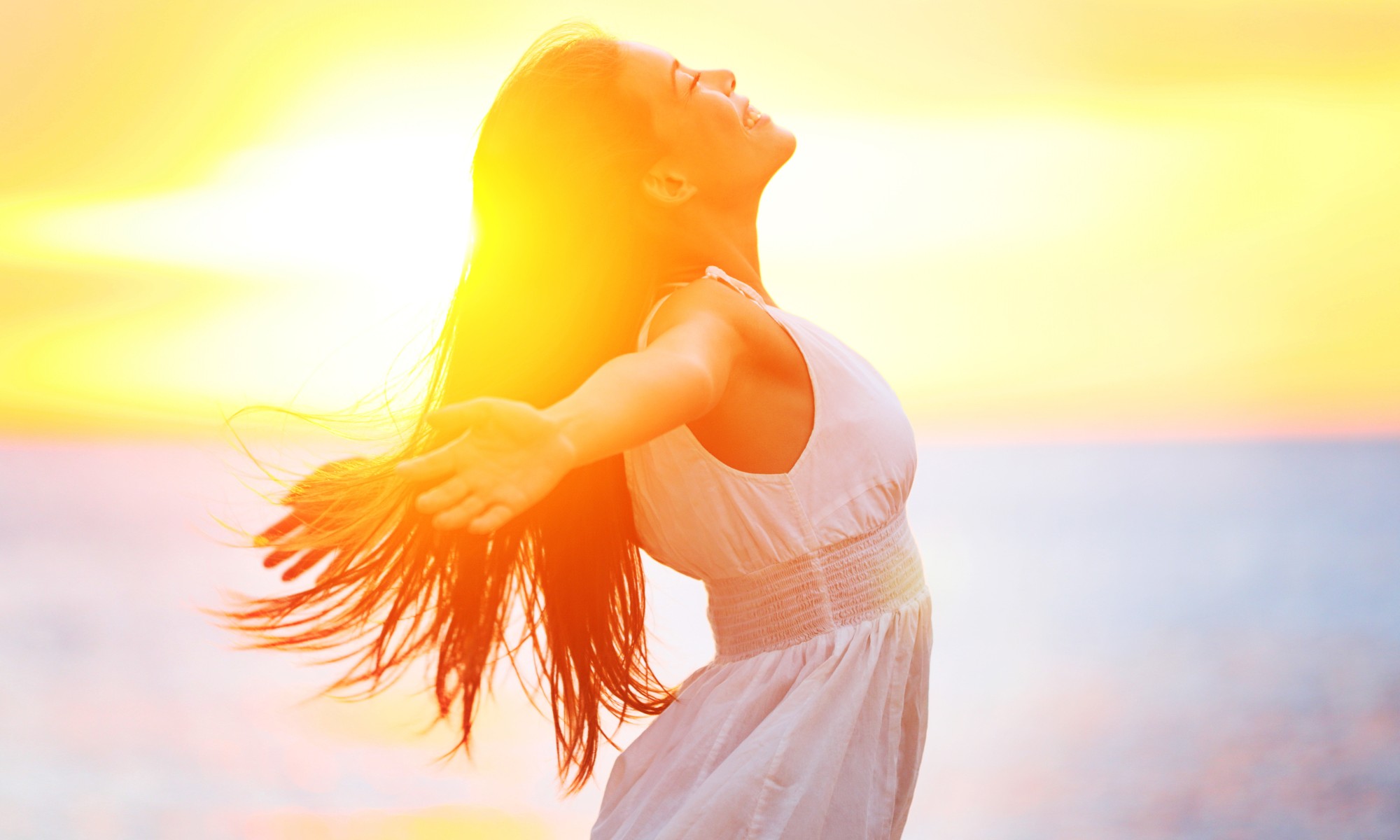 Fotografia de uma mulher de braços abertos para a vida, sorrindo, de olhos fechados e direcionada ao céu. A fotografia é iluminada pela luz do sol. Simboliza o ato de colocar a si mesma em primeiro lugar e seguir seus próprios desejos, abrindo mão da necessidade de cumprir as expectativas dos outros.